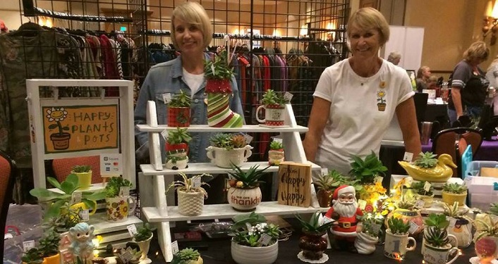 Two friendly looking craft vendors standing behind their merchandise table. 