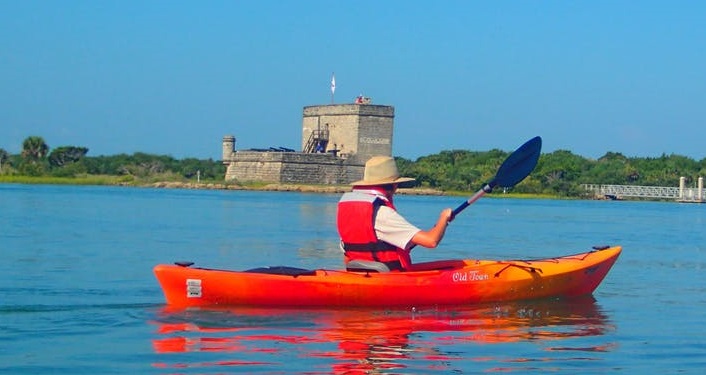 image of bright orange kayak with person wearing white shirt and life vest, paddling in front of Fort Matanzas on a Fort Matanzas Kayak Trip