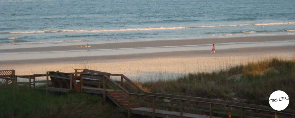 Image contains the outdoors, sand, marsh, a boardwalk, the ocean, and two people.