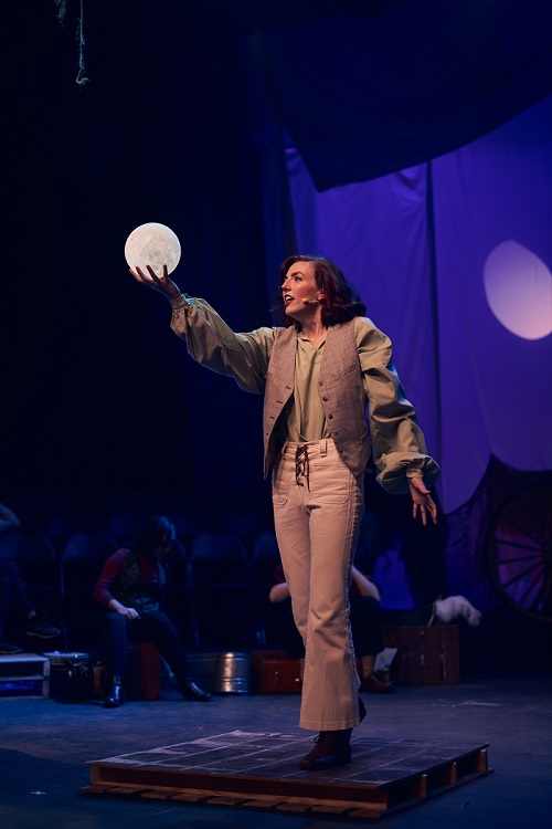 Image contains a woman standing straight holding a fake moon in her hand.