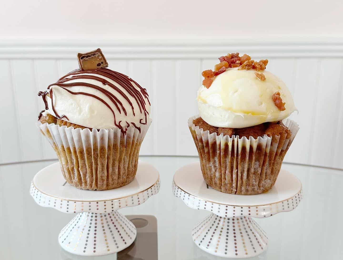 Two cupcakes on little pedestals with chocolate and bacon crumble toppings.