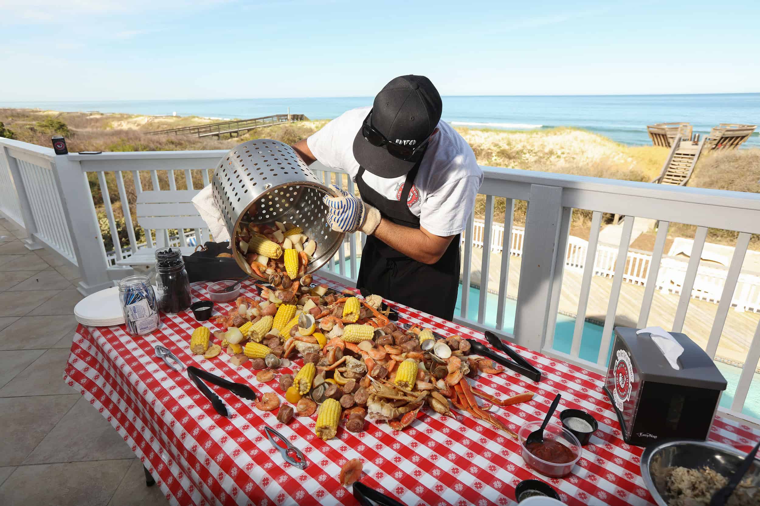 A man in a black apron and hat standing on a balcony with the beach in the background pouring out a seafood boil (crab legs, crawfish, corn on the cob) out onto a red and white checkered table cloth.