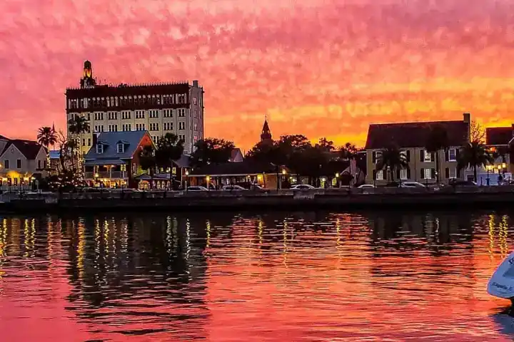 A view of St. Augustine buildings from the river set against a pink and soft orange sky at sunset, which is reflected in the calm water.