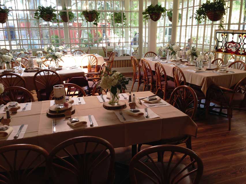 Interior view of Raintree Restaurant's dinning room. Bay windows line the room, potted plants hang from the ceiling, and large, rectangular tables are adorned with candles, silverware, and vases full of flowers.