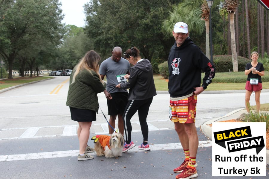 Runners getting ready for the Run-off-the-turkey run. Two runners are helping another pin on his paper number. One of the runners also has a small long-haired dog dressed in a turkey costume.