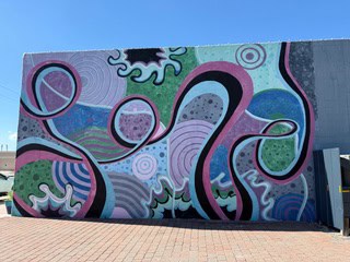 A wall mural on the exterior of a building. Shades of blue and pink, and green dominate the wall in the form of flowing curvy lines and semi-circular shapes. 