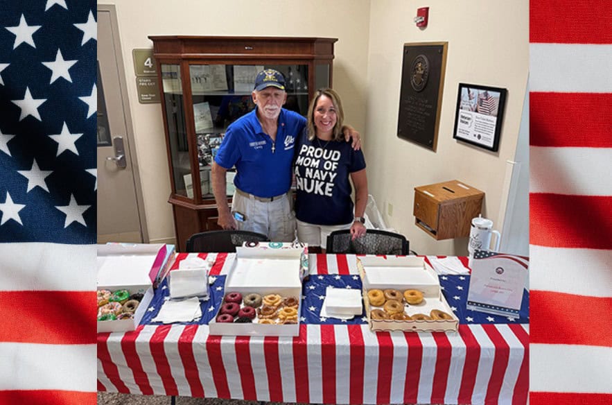 A veteran with his arm around his daughter. They are both standing behind a table with an American flag themed table cloth. On top of the table are boxes of doughnuts. 