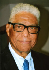 Photo of Dr. Robert Hayling, a brown skinned man with grey hair, sporting rectangular black-framed glasses. 